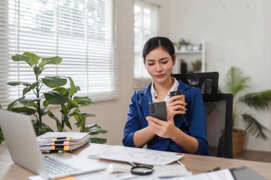 Profesyonel bir kadın elinde kahve fincanı tutarken akıllı telefonuyla meşgul olur, iş ve rahatlama karışımı sergiler..