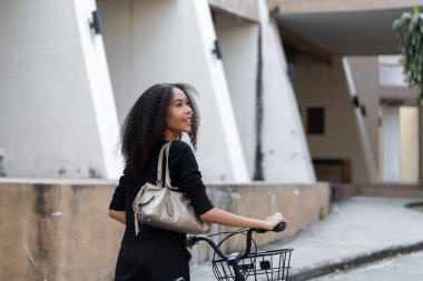 Omzuna takılı şık bir el çantasıyla, genç bir kadın bisiklet sürerken arkasına bakıyor, kentsel sürdürülebilirlik ve toplum ruhunu somutlaştırıyor..