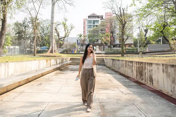 Bir kadın şehir parkında yürümekten zevk alır, yeniden kullanılabilir fincan ve çantasını güvenle tutar, sürdürülebilir bir yaşam tarzı ve pozitifliği somutlaştırır..