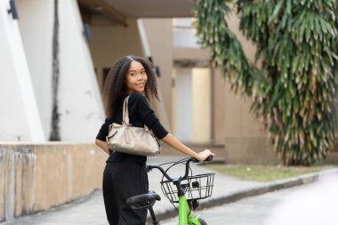 Kentsel bir arka planda, bir kadın bisikletini gülümseyerek sürüyor, kolaylıkla stili, sürdürülebilirliği ve sağlıklı yaşamı birleştiriyor..