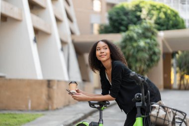 Sürdürülebilir yaşam hedeflerine odaklanan bu kadın, şehir zeminindeki canlı yeşil bisikletine yaslanıp, şehriyle bağlantı kurarak kahve molası veriyor..