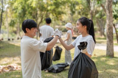 Bir grup genç gönüllü parkta çöp toplarken gülümsüyor, takım çalışmasını ve toplumsal müdahaleyi vurguluyor.