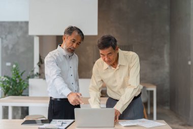 Çağdaş bir ofis ortamında, üst düzey bir yönetici genç bir meslektaşıyla görüşür, önemli iş stratejilerini tartışır ve dizüstü bilgisayardaki verileri inceler.,