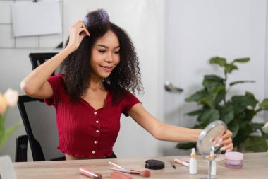 Buklelerini kucaklayan bir kadın, hayat dolu bir ev ortamında neşe ve kendini beğenmişliğini ifade ederek güzellik ürünleriyle meşgul olur..