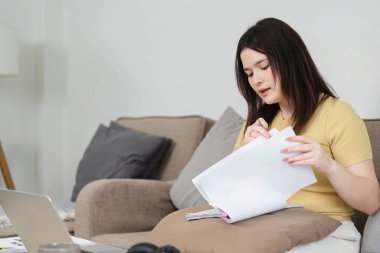 Bu resim, evdeki iş belgelerine odaklanan, uzaktan çalışma oturumlarında organizasyon ve önceliklerin önemini vurgulayan bir kadını resmediyor..