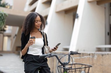 Sıcak bir şehir ortamında, bir kadın kahvesini yudumlamak ve telefonunu kontrol etmek için bir dakikasını ayırarak günlük işe gidip gelmeye modern ama rahat bir yaklaşım sergiliyor..