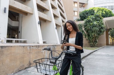 Tarz sahibi genç bir kadın bisikletinin yanında akıllı telefonuyla meşgul oluyor, sürdürülebilir bir şehir yaşam tarzı ve modern bağlantıyı somutlaştırıyor, çevre dostu ulaşımı teşvik ediyor..