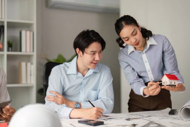 Mimari planlar üzerinde işbirliği yaparlarken, ekip ayrıntıları coşkuyla tartışarak, model bir evin etrafında toplanan üretken bir fikir alışverişini kolaylaştırıyor..