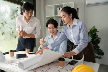 Üç meslektaş bir tasarım oturumuna katılıyor, bina planları ve taslaklarını inceliyor, işbirliği ve yenilikçi düşünce sergiliyor.