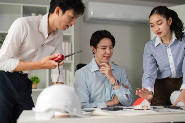 Üç meslektaş, küçük bir model ev ve planları incelerken proje detaylarını tartışarak dinamik bir beyin fırtınası yapıyorlar. Atmosfer işbirlikçi ve yeniliğe odaklı..