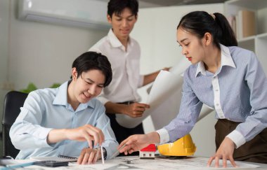 İşbirlikçi bir çalışma alanında, ekip üyeleri bir model ev ve planlarla uğraşırken kendi tasarım fikirlerini ve inşaat stratejilerini tartışarak yenilikleri teşvik ediyorlar..