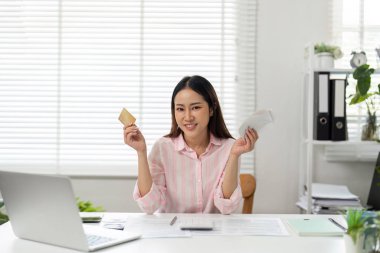 Parlak bir ev ofisinde, bir kadın mutlu bir şekilde elinde kredi kartı ve makbuzlarla poz verir, kişisel finans yönetiminde başarı ve yetkilendirmeyi temsil eder..
