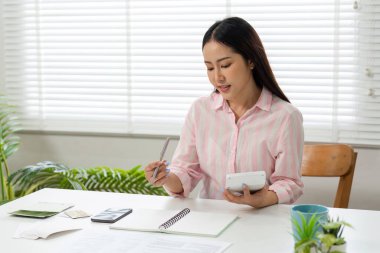 Bütçesine odaklanan bir kadın, hesap makinesinden faydalanıyor ve finansmanını parlak bir ofis alanında yönetirken notlar alıyor..