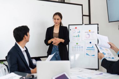 Hareketli bir ofis ortamında, genç bir kadın önemli bütçe anlayışları sunarken, meslektaşları bu bilgiyi özümserek iş dünyasındaki etkili iletişimi vurguluyor..