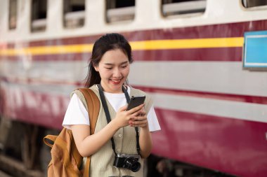 Tren istasyonunda neşeli bir kadın duruyor, akıllı telefonunu kontrol ederken gülümsüyor, kamerasıyla seyahat yolculuğunu belgelemek için heyecanlı..
