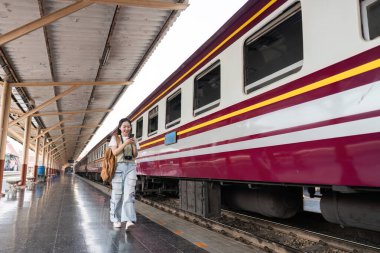 Genç bir kadın platformda yürüyor, telefonu elinde ve yüzünde kararlı bir gülümseme, seyahat yolculuğuna çıkmaya hazır..