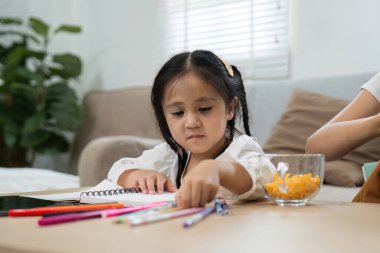 Genç bir kız çalışma alanını canlı renkli kalemler ve bir kase abur cuburla kuruyor. Evdeki öğrenme rutinine eğlenceli ama odaklanmış bir yaklaşım sergiliyor..