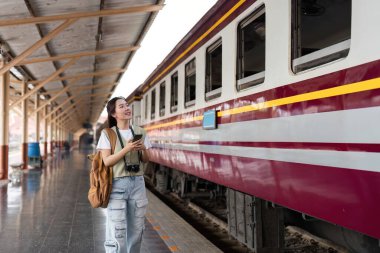 Yanında akıllı telefonuyla, genç bir kadın düşünceli bir şekilde tren istasyonuna bakıyor, yolculuğunun tadını çıkarıyor..