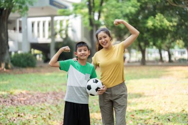 Bir anne ve oğlu aktif yaşam tarzlarını bir futbol topuyla gururla poz vererek kutluyorlar. Gülüşleri neşe ve coşkuyu yansıtıyor. Fiziksel sağlık ve aile bağları için..