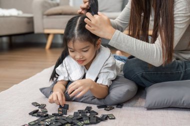 Yerde domino taşlarını düzenlerken, bir anne kızının saçlarını taramak için biraz zaman ayırıyor, günlük yaşamlarında bir neşe ve şefkat karışımı sergiliyor..