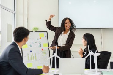 Parlak bir ofis alanında, bir kadın görsel araçlar kullanarak ESG stratejilerini etkili bir şekilde iletir ve meslektaşlarını sürdürülebilirlik diyaloğuna aktif olarak katılmaya teşvik eder..