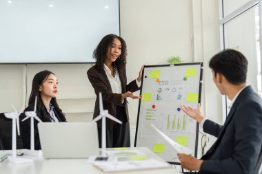 Dinamik bir atmosferde, bir kadın ESG girişimleri hakkında çarpıcı bir tartışma yürütüyor, ekibi dikkatle dinlerken ve katılımcılar arasında önemli performans göstergeleri sergiliyor..