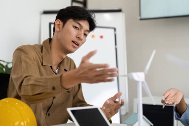 İşbirlikçi bir çalışma alanında, genç bir profesyonel, sürdürülebilir tasarımda yenilik sinyalleri veren minyatür bir rüzgar türbini modelini işaret ederken rüzgar enerjisinin faydalarını anlatıyor..