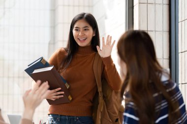 Elinde kitaplarla, bir öğrenci neşeli bir şekilde arkadaşlarına el sallıyor, heyecan ve dostluk duygusunu canlı bir ortamda ifade ediyor..