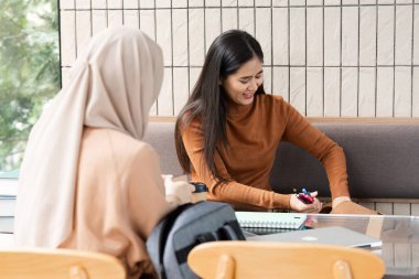 İki öğrenci bir kafede çalışmalarına odaklanmış, sınavlara hazırlanırken notlarını ve fikirlerini paylaşıyor, takım çalışmasının ve akademik işbirliğinin özünü vurguluyor.
