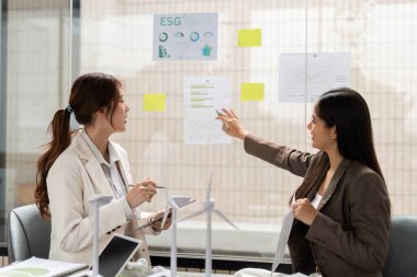 İki meslektaş ESG kavrayışları hakkında canlı bir tartışmaya giriştiler, duvardaki çizelgelere işaret ederek, sürdürülebilir uygulamaları iş modellerine entegre etmeye olan bağlılıklarını gösterdiler..