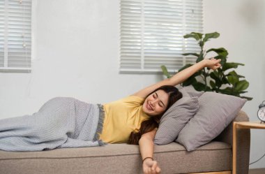 Genç bir kadın kollarını parlak bir gülümseme ile uzatıyor, oturma odasında sabah güneşinin tadını çıkarıyor. Bu an, güne olumlu başlamanın mutluluğunu yakalar..