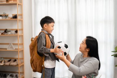 Bir anne okula hazır olan oğluna neşeyle bir futbol topu verir. Bu an, çocukların hayatındaki akademik ve spor dengesini yansıtıyor..