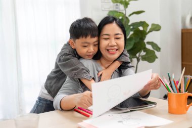 Bir anne ve oğlu, ona arkadan sarılırken aynı sevgi dolu anı paylaşıyorlar. İkisi de onun yaratıcı çiziminden memnunlar ve yakın ilişkilerini gösteriyorlar..