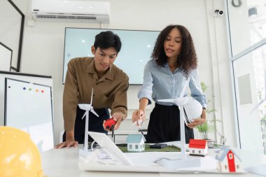 İki genç profesyonel, modern ofis ortamında sürdürülebilir konut çözümleri, rüzgar türbinleri ve model evler sergilenmesi konusunda işbirliği yapmaktadır..