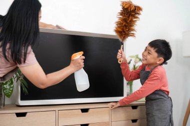 Bir anne, oğlu televizyon standının tozunu alırken temiz sprey sıkar. Evlerinde neşeli ve etkileşimli bir temizlik deneyimi yaratır..