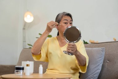 Son sınıf öğrencisi bir kadın cilt bakımından zevk alır. Yansımasına bakarken serumu dikkatle uygular. Bu an, yaş çeşitliliğini ve kendine bakımı vurguluyor.
