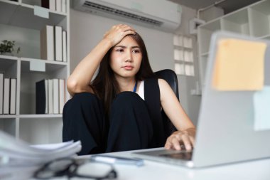 Genç bir iş kadını, modern bir ofis ortamında masasında çalışırken yorgun ve stresli görünür..