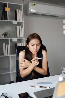 Bir iş kadını çalışma masasında kendini iyi hissetmiyor, termometreyi tutuyor, modern ofis ortamında iş için gerekli malzemelerle çevrili..