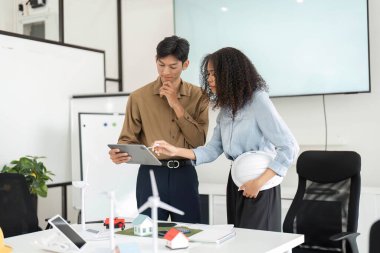 Bir erkek ve bir kadın sürdürülebilirlik hakkında verimli bir tartışmaya girişiyor, çevresi çevre dostu modeller ve malzemelerle çevriliyken tablet üzerindeki planları gözden geçiriyorlar..