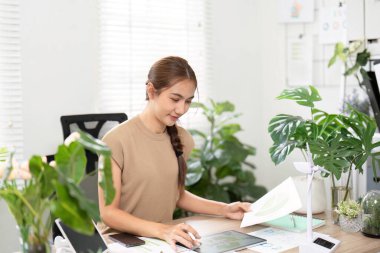 Genç bir kadın, çevre dostu bir ofis alanında yeşil alanlarla çevrili tablet ve notlarını kullanarak sürdürülebilir iş uygulamaları yapıyor..