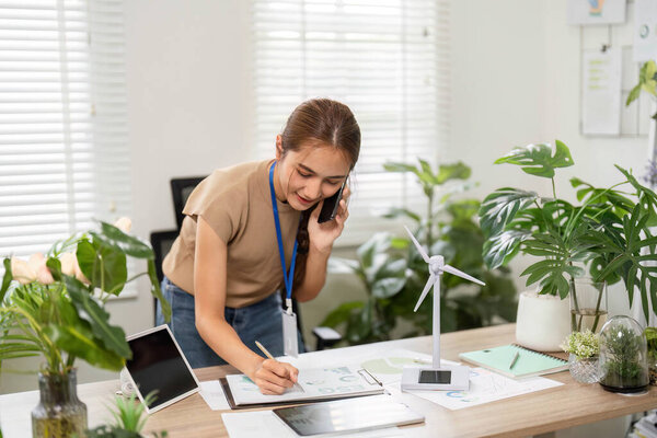 Сфокусированная молодая женщина записывает заметки во время телефонного звонка об экологических инициативах в ярком офисе, наполненном зеленью.