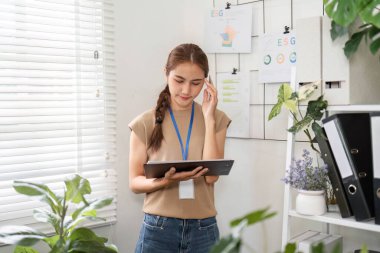 Telefonda ESG stratejilerini tartışan genç bir kadın notlarını yeni ve modern bir ofiste gözden geçirip sürdürülebilirliği savunuyor..