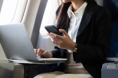 Genç bir kadın, iş seyahatinde birden fazla görevi vurgulayarak, bir uçakta aynı anda akıllı telefonunu ve laptopunu kullanıyor..