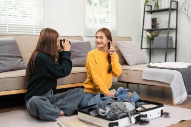 İki arkadaş, bavullarını toplarken neşeli anları yakalıyor, gülümsüyor ve rahat bir ev ortamında poz veriyorlar..