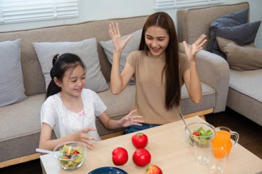 Neşeli bir anne ve kız, salata ve meyve suyu da dahil olmak üzere sağlıklı yemekleri paylaşıyor, evde keyifli bir yemek deneyimi sırasında sağlık ve aile bağlarını vurguluyor..