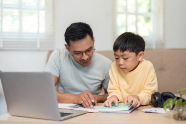 Bir baba, oğlunun okul seansında ders çalışmasına yardımcı oluyor. Her ikisi de not defterlerine odaklanıyor..