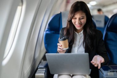 Gülümseyen bir iş kadını, uçarken laptopuyla kahve yudumlarken modern iş seyahatinin keyfini yansıtıyor..