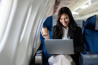 Uzaktan kumandayla çalışan profesyonel bir kadın uçağın içinde iş seyahatinin özünü kahveyi yudumlarken gülümseyerek somutlaştırıyor..