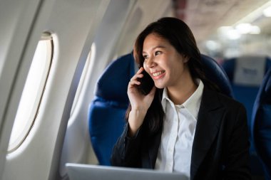 Bir uçağın içinde telefonuyla konuşan neşeli bir kadın iş seyahatlerinin dinamik doğasını ve uçuşlar sırasında uzaktan bağlantıları simgeliyor..