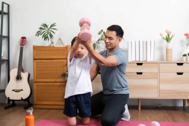 Bir baba, bir dambılı kaldırırken oğluna yardım eder, spor yapmayı teşvik eder ve eğlenceli bir ev ortamında güç geliştirmeyi teşvik eder..